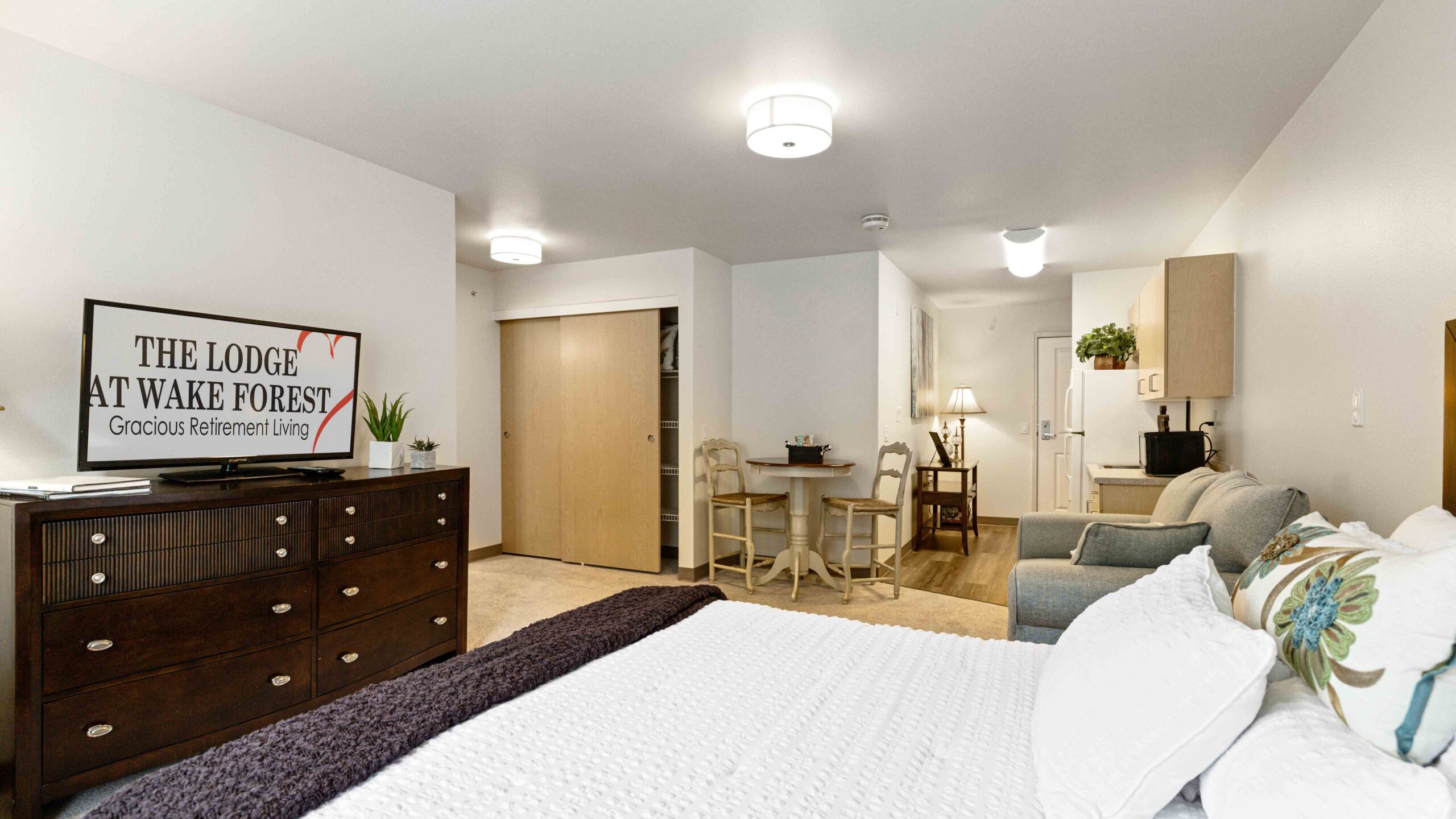 A bright, modern studio apartment with a bed, dresser, TV, small dining table, kitchenette, and a cozy sitting area. The decor is neutral with white walls and light wood furniture.