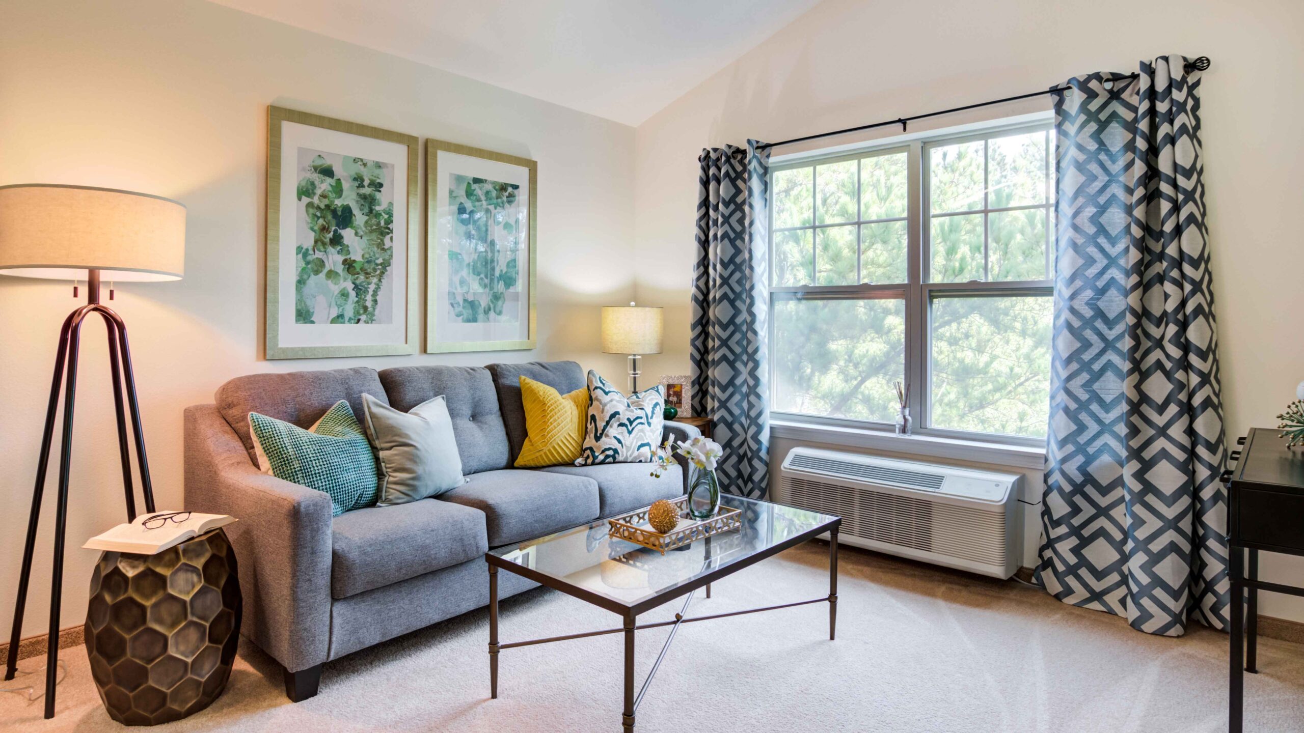 A cozy living room with a gray sofa, colorful pillows, two abstract art pieces on the wall, a glass coffee table, two lamps, patterned curtains, and a large window letting in natural light.