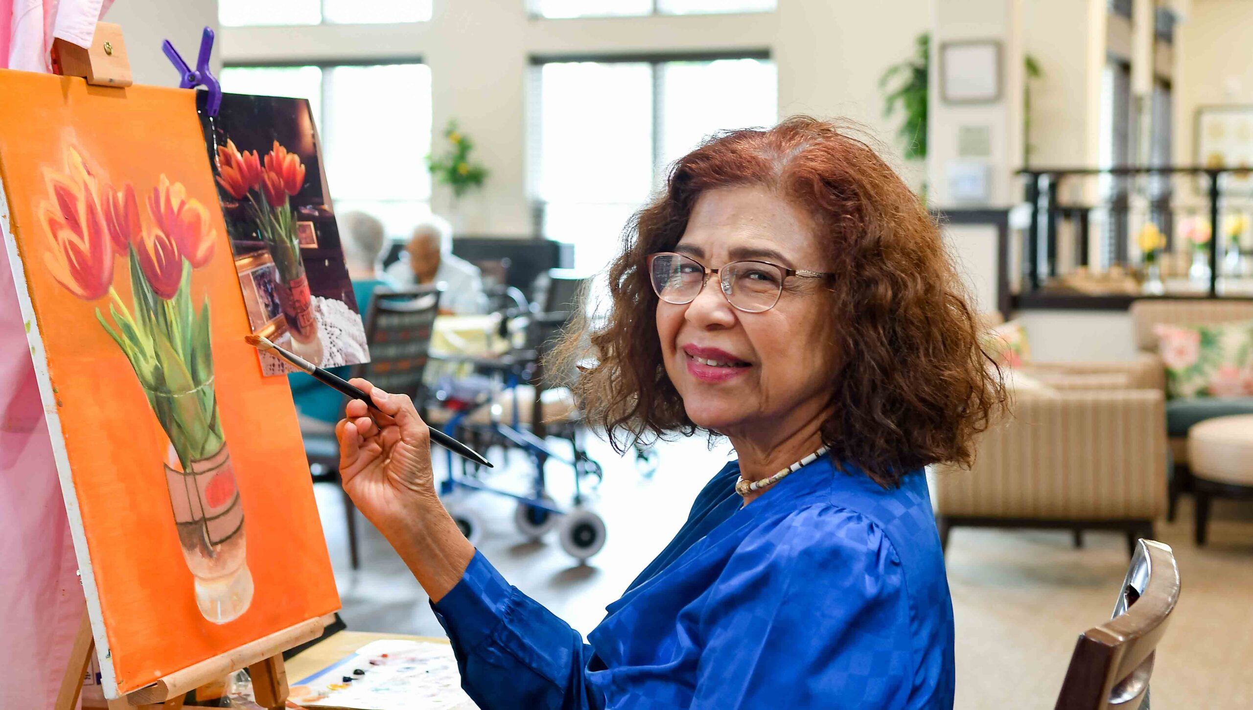 An elderly woman wearing glasses and a blue blouse smiles while painting red and orange tulips on a canvas in a bright, cozy room. She holds a paintbrush and looks toward the camera.