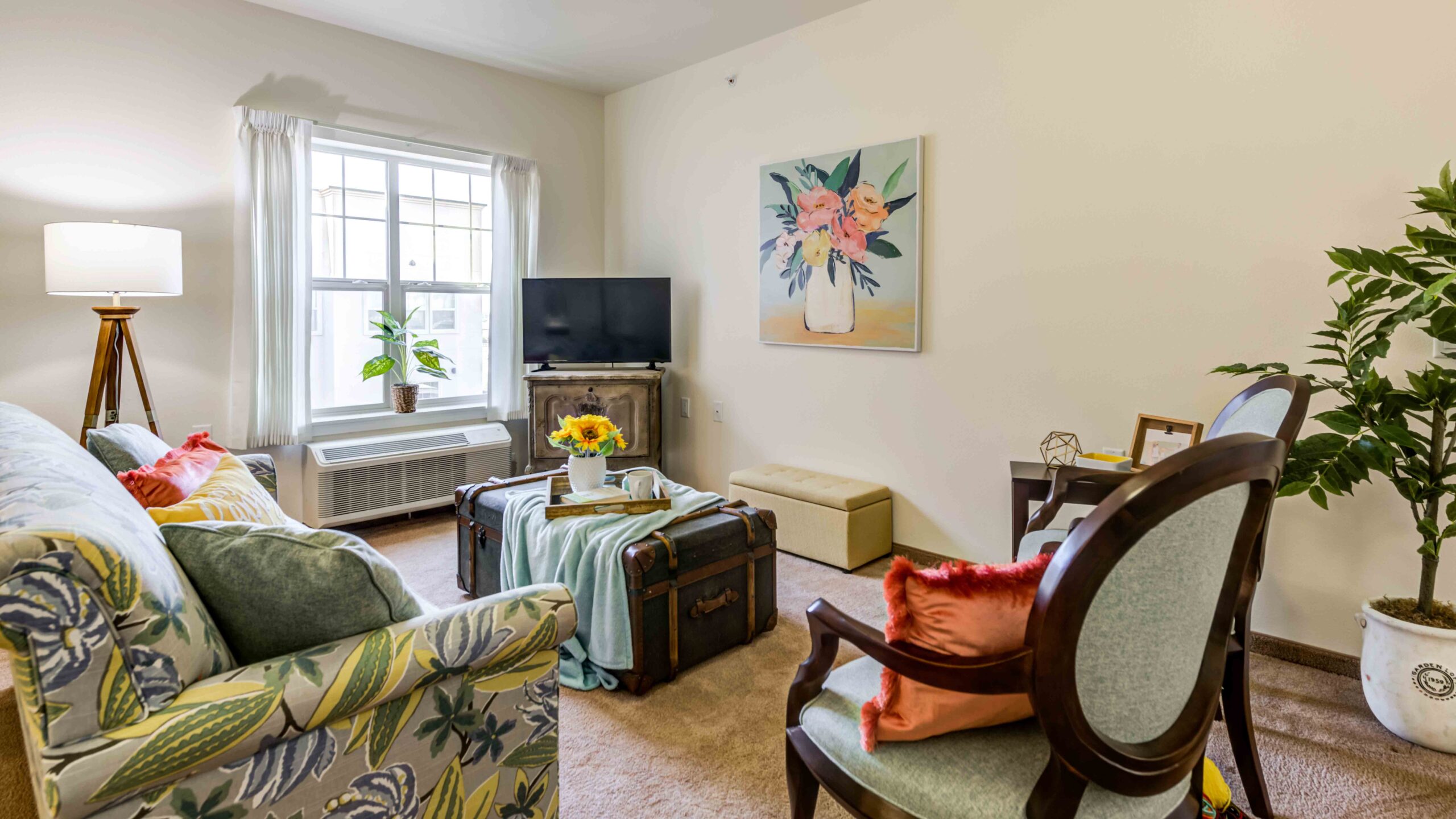 A cozy, well-lit living room with a floral sofa, two chairs, a trunk as a coffee table, a TV on a stand, a potted plant, a large window with white curtains, and a floral painting on the wall.