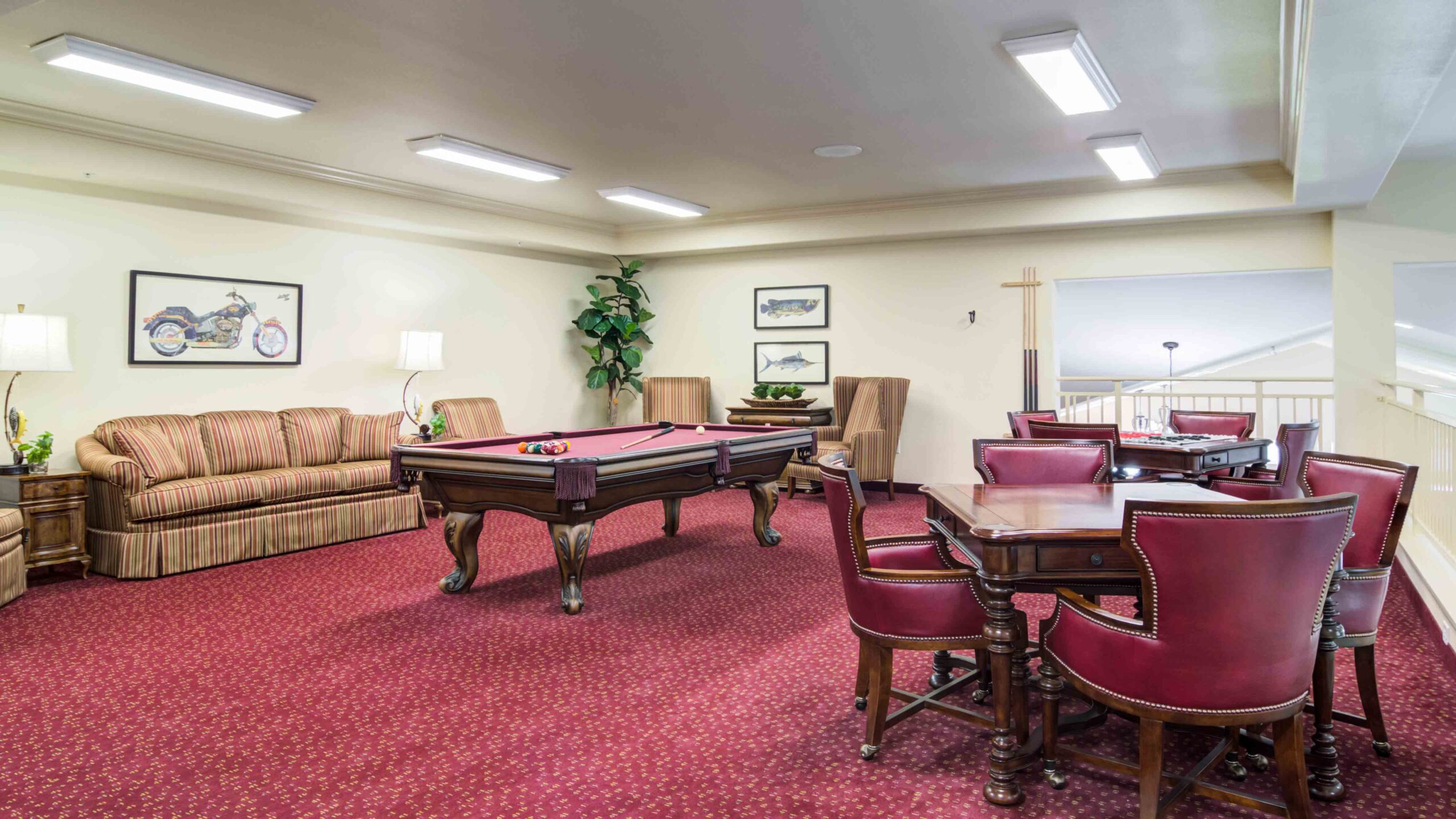 A game room with a red pool table, red upholstered chairs around a card table, a striped sofa, framed artwork on cream walls, and red carpet flooring. Plants and lamps are placed around the room.