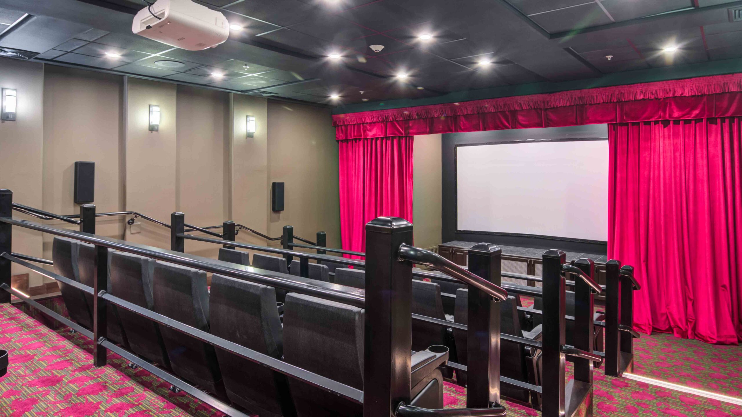 A small movie theater with black seats, red patterned carpet, a large white screen framed by red curtains, and wall-mounted lights, viewed from the back of the room.