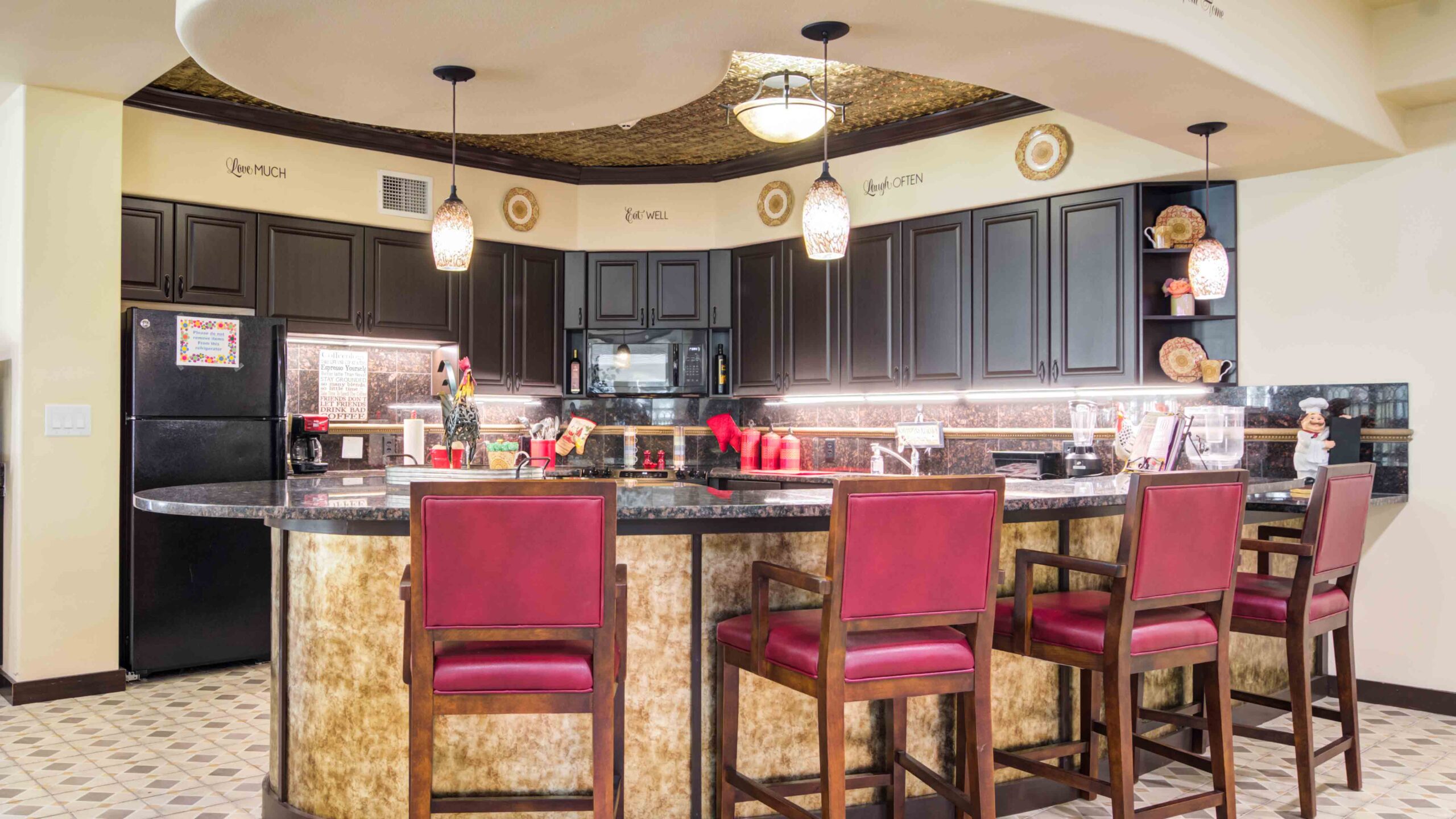 A modern kitchen with dark cabinets, a black refrigerator, and microwave, a curved granite countertop with four red-cushioned bar stools, pendant lights, and decorative wall accents.