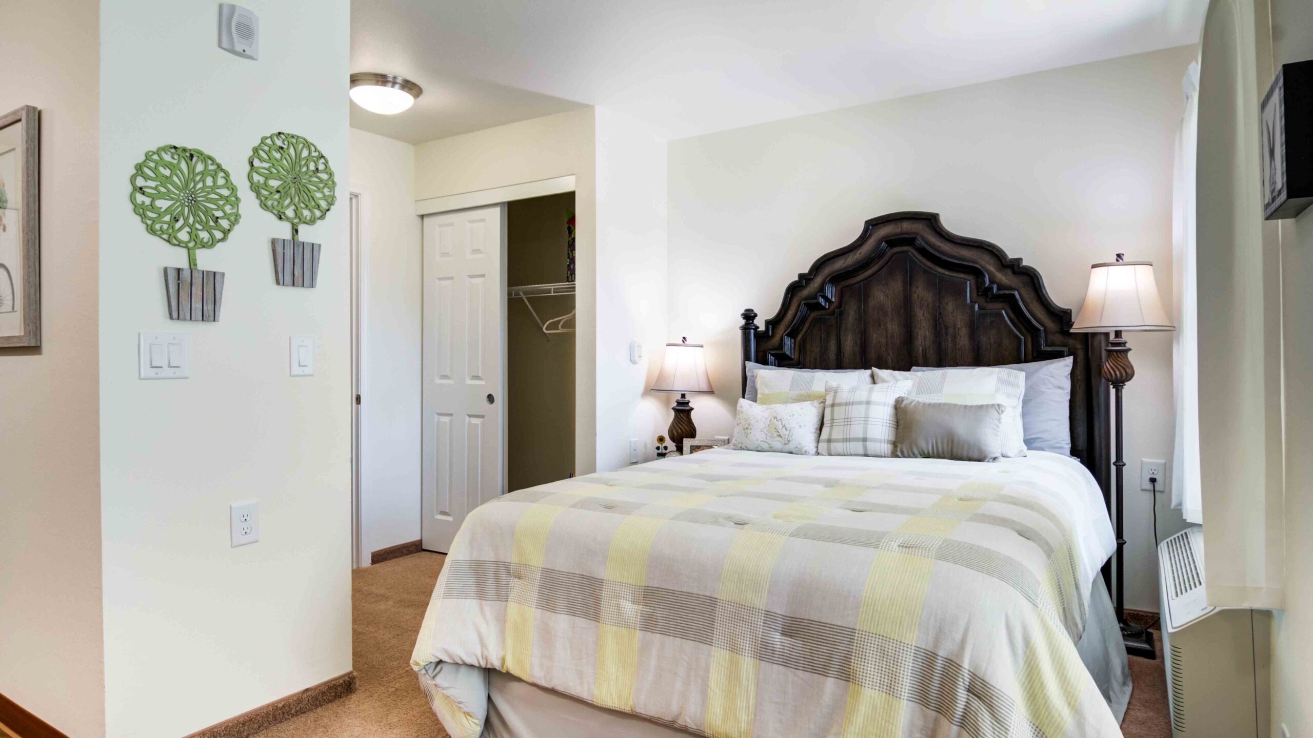 A cozy bedroom with a dark wooden headboard, neatly made bed with light-striped bedding, two lamps on bedside tables, wall art of two green trees, and an open closet door.
