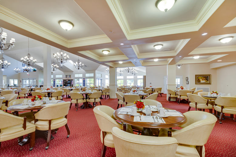 A spacious, well-lit restaurant dining room with red carpet, cream-colored chairs, and round wooden tables set for dining. Chandeliers hang from the ceiling, and large windows allow natural light to brighten the room. Small flower centerpieces adorn the tables.