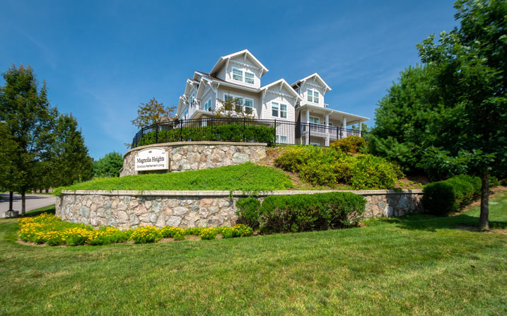 A large, white, two-story house with multiple balconies sits on a sloped, landscaped hill, surrounded by lush greenery and trees. The hill features a stone retaining wall and a sign that reads 