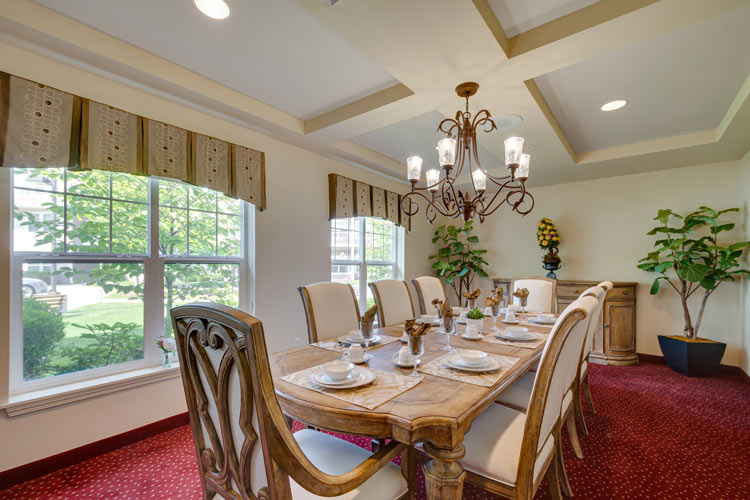 A bright dining room with a long wooden table surrounded by eight upholstered chairs on a red carpet. The table is set for a meal, and a chandelier with multiple lights hangs from the ceiling. There are two large windows, and potted plants are placed around the room.