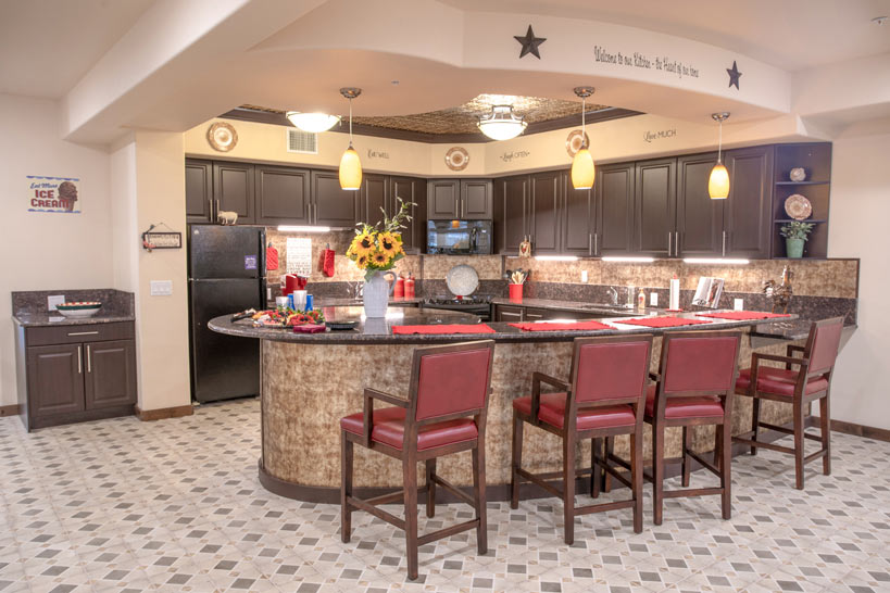 A spacious kitchen with a curved island featuring four high-backed chairs with red cushions. The countertops are black, and the cabinets are dark wood. Sunflowers in a vase sit on the island. The tiled floor has a geometric pattern, and various decorative signs adorn the walls.