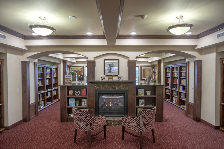 A cozy library with red carpeted flooring, featuring two patterned chairs in front of a fireplace. Bookshelves filled with books line the walls, and warmly lit ceiling lights create a welcoming atmosphere. Decor includes framed art and plants on the mantel.