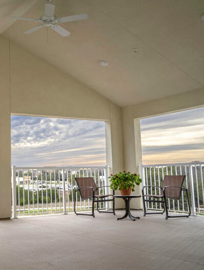 A spacious balcony with two brown chairs and a small round table with a potted plant. The balcony has a white railing, and it overlooks a landscape with buildings and greenery under a cloudy sky. A white ceiling fan is mounted above, offering a serene viewing spot.