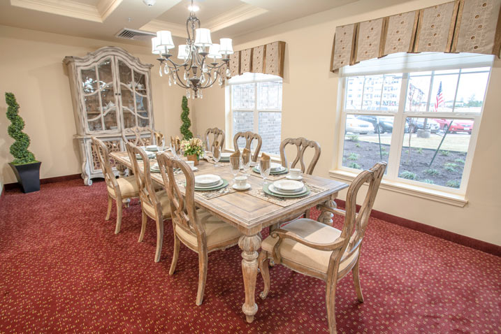 A dining room with a large wooden table surrounded by eight elegant chairs. Table settings include plates, glasses, and cutlery. A chandelier hangs above the table. A cabinet with decorative dishes is against the wall, and windows let in natural light.