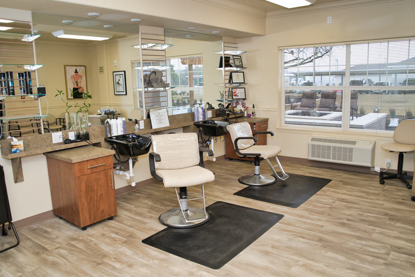 A clean, modern hair salon with two styling stations featuring mirrors, chairs, and sinks. The room has wood flooring and large windows letting in natural light. Shelves hold various hair products and framed pictures decorate the walls.