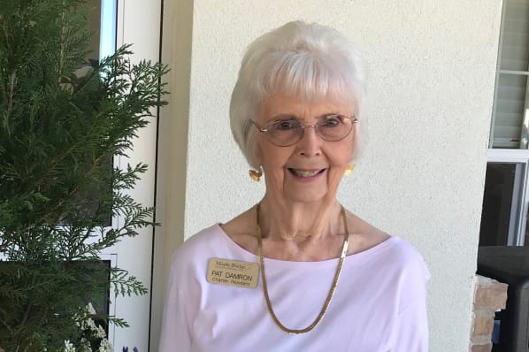 An elderly woman with short white hair and glasses smiles while standing outside near a plant. She is wearing a light pink top, gold earrings, a matching necklace, and a name tag.
