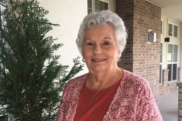 An elderly woman with short, curly gray hair, wearing a red patterned shawl and a red top, stands outside near a brick building. She is smiling and has a gold necklace. A green bush is visible next to her.
