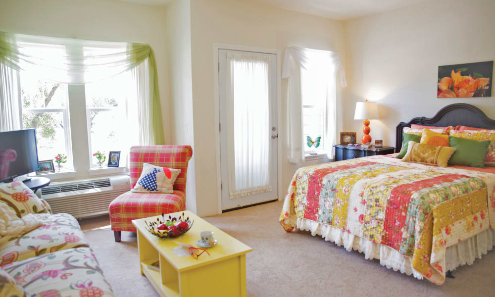 A cozy bedroom features a large colorful quilt-covered bed, a plaid armchair, and a small yellow coffee table. A window with green curtains and a glass door with white drapes allow natural light in. Decor includes a framed orange flower painting and various ornaments.