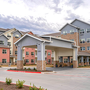 A modern, multi-story building with pastel-colored walls and large windows, featuring an entrance canopy supported by stone pillars. The surrounding area includes well-maintained landscaping and a paved driveway under an overcast sky.