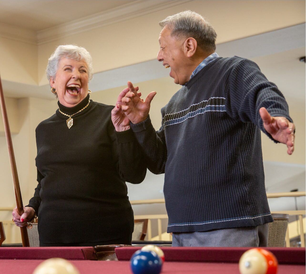 Two elderly individuals are standing near a billiards table, laughing joyfully. One person, holding a pool cue, is dressed in a black sweater with a necklace, while the other, wearing a dark striped sweater, gestures animatedly with an open hand. Several pool balls are visible.