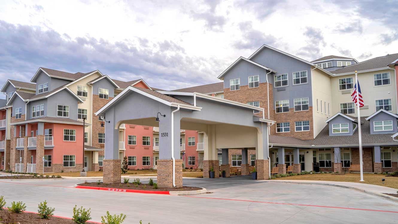 A multi-story building complex with a mix of peach, beige, and gray facades. It features balconies and a covered entrance with pillars. The surroundings include a neatly paved driveway, small shrubs, and an American flag on a flagpole in front.