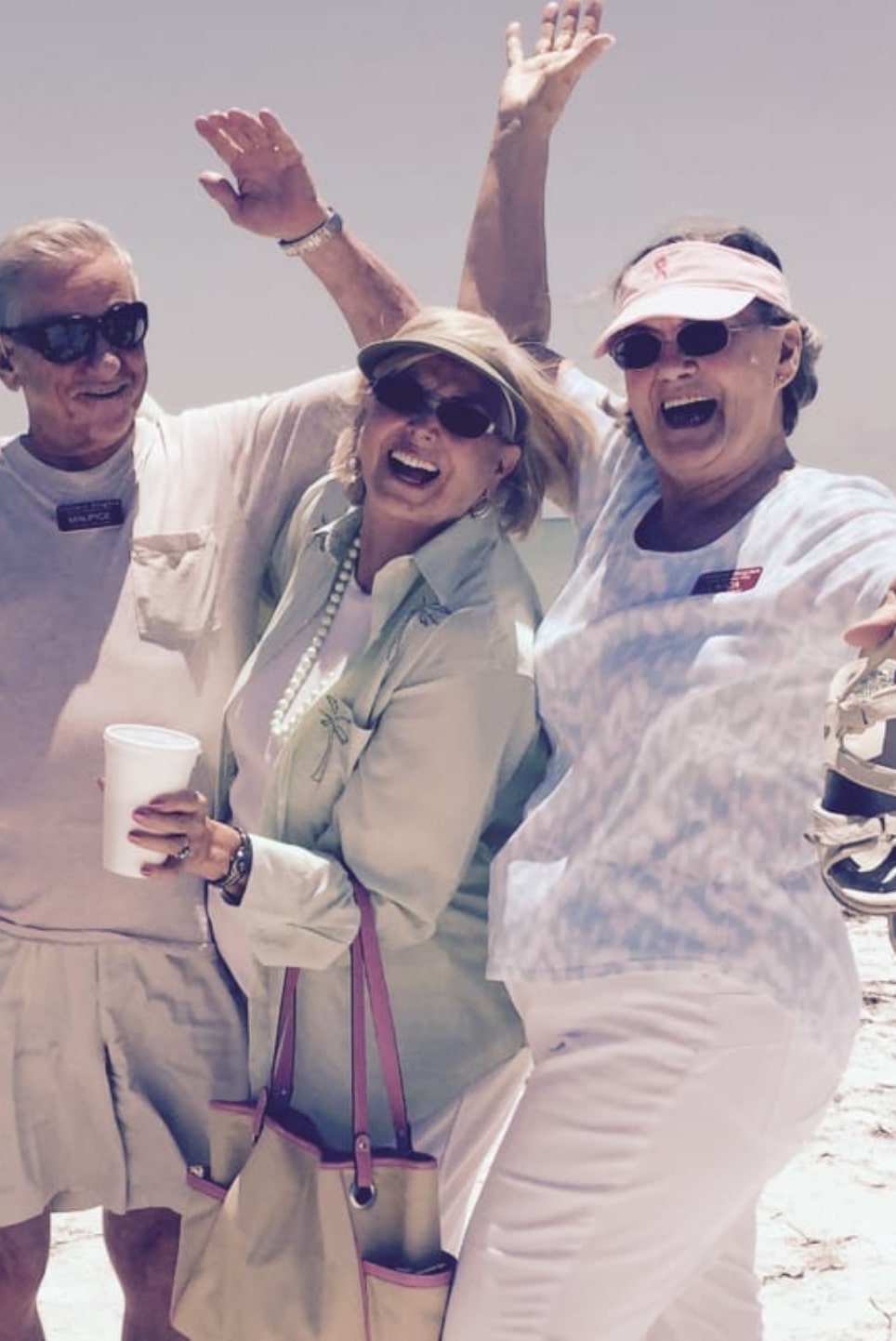 Three people joyfully pose with wide smiles and raised arms on a sunny day at the beach. One man and two women are dressed in light, summery clothing, with one woman wearing a sun visor and another carrying a pink-trimmed bag. The beach and ocean are visible in the background.