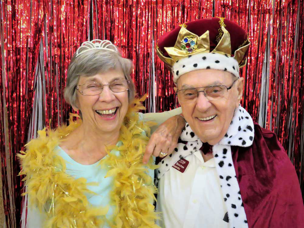 Two elderly individuals smiling in front of a red tinsel backdrop. The person on the left is wearing glasses, a tiara, and a yellow feather boa. The person on the right is wearing glasses, a gold and maroon crown, and a red robe with white and black fur trim.