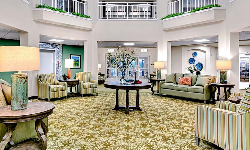 A spacious living area with green-patterned carpet, several armchairs, a sofa, and a central table. Lamps with beige shades are on side tables. Walls are decorated with paintings and floral arrangements, and a balcony overlooks the room from above.