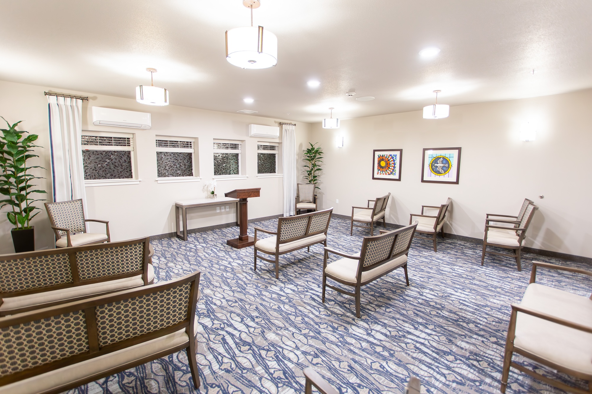 A room with chairs arranged in a semi-circle around a wooden podium. The floor is covered with a blue and white patterned carpet. The walls are adorned with framed artworks, and tall plants are placed in the corners. Ceiling lights illuminate the space.