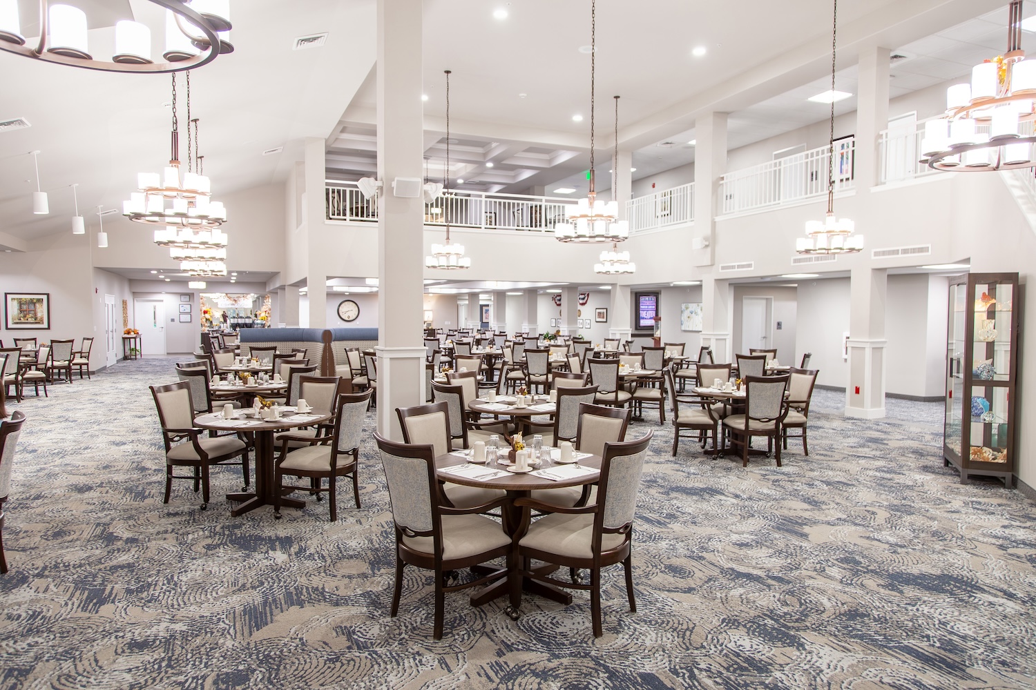 A spacious dining room with multiple tables set for a meal, featuring elegant light fixtures and a patterned carpet. The room has high ceilings and a mezzanine area visible, creating an open and airy atmosphere.