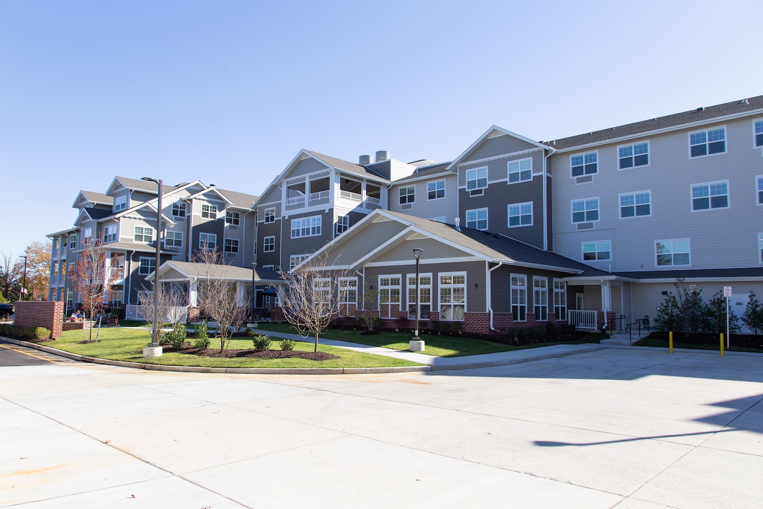 A modern apartment complex with multiple stories, featuring a gray facade and large windows. The building is surrounded by neatly landscaped greenery and a clear, blue sky.