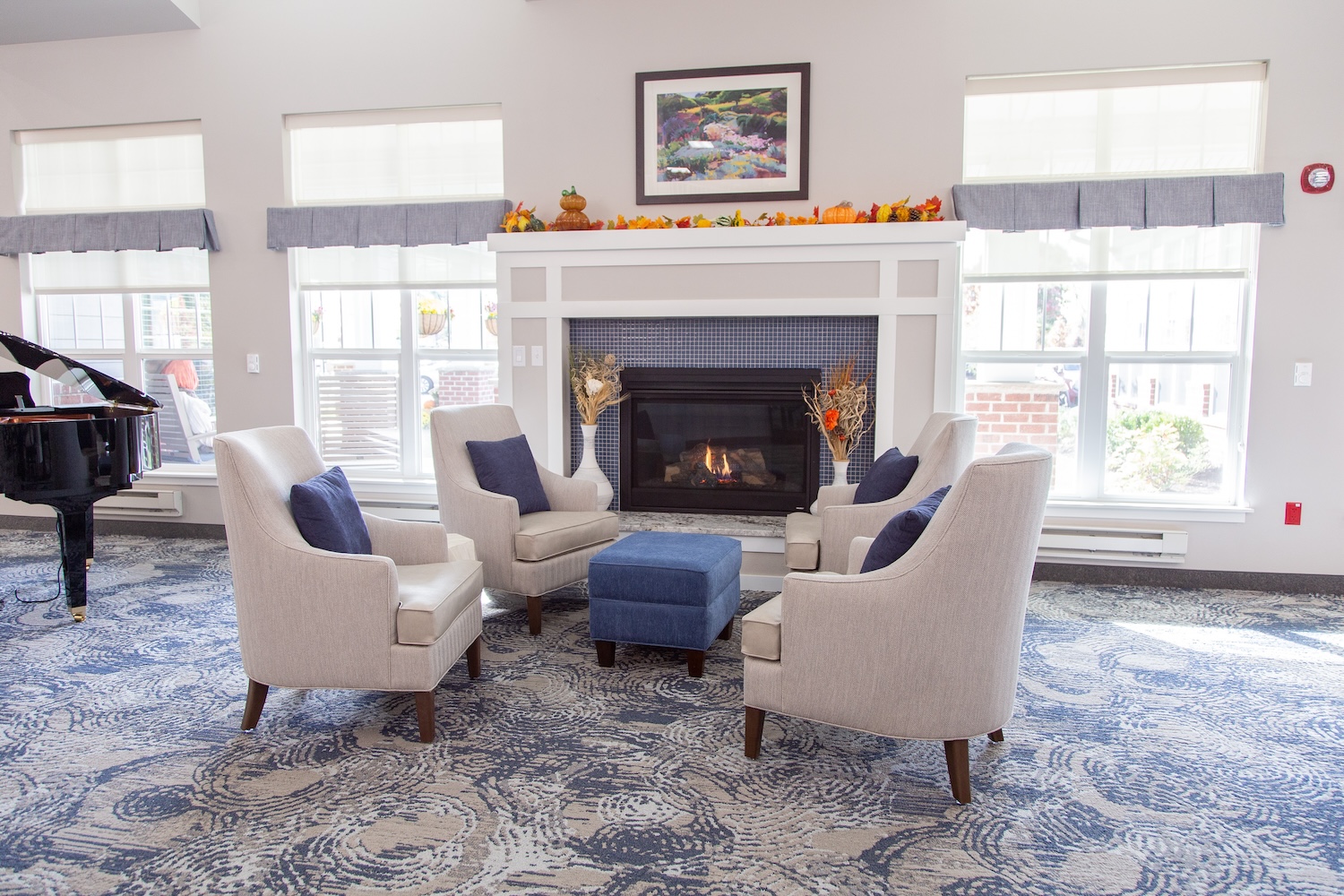 A cozy living room features a fireplace with autumn decor, four beige armchairs, and a blue ottoman. Large windows with white blinds brighten the space, and a black grand piano is visible on the left side.