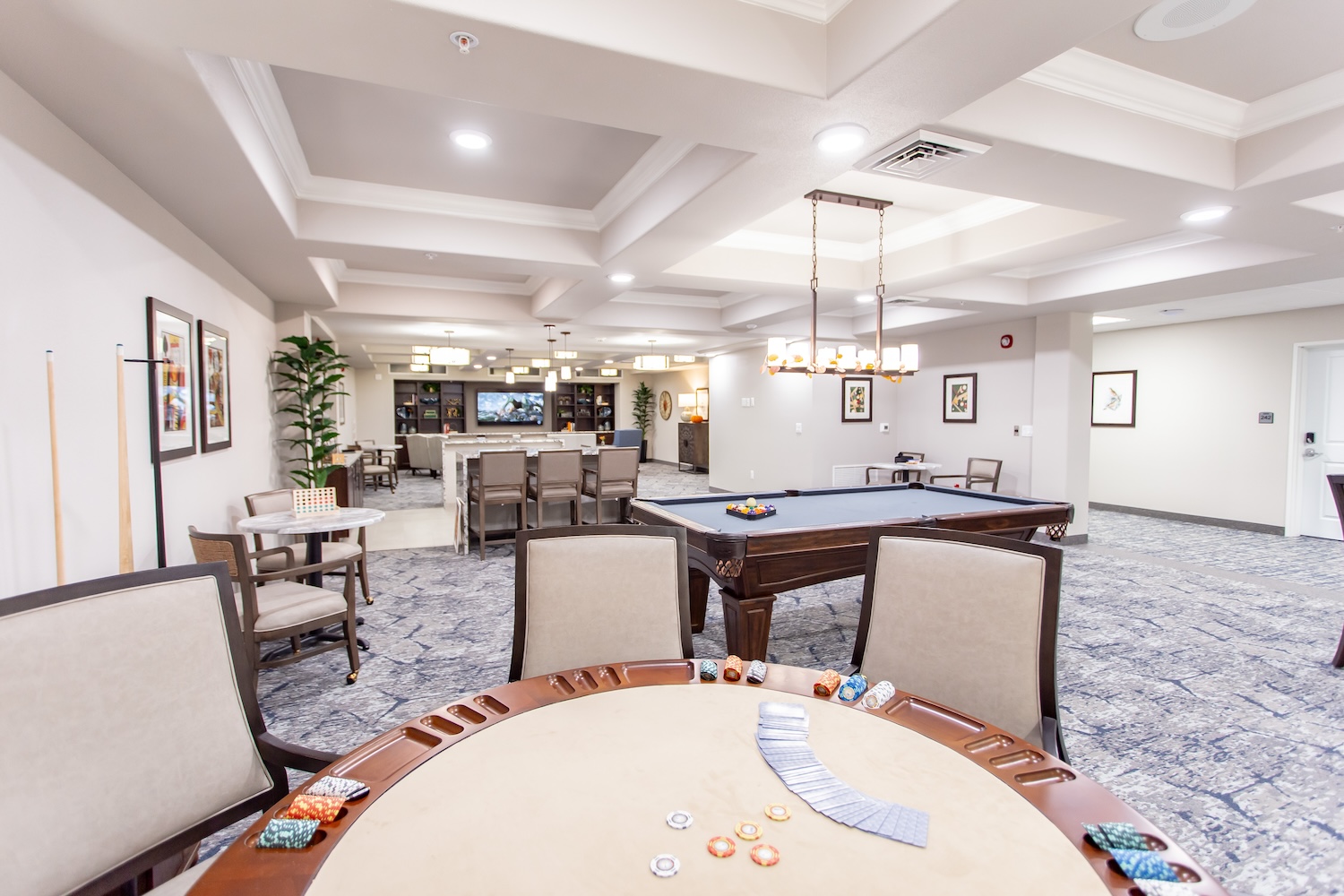 A recreation room with a poker table in the foreground, a pool table in the center, and a bar area at the back. The room is well-lit with ceiling lights and decorated with framed art and plants. Chairs are arranged around the tables.