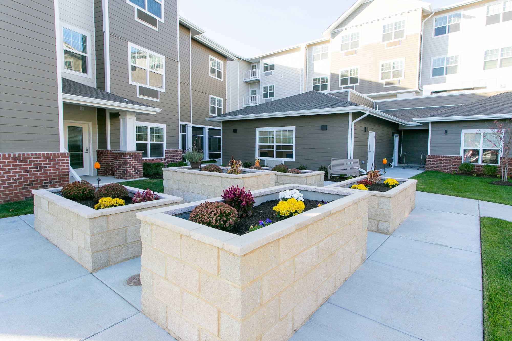 A modern apartment complex courtyard featuring raised stone planters with blooming flowers. The buildings are three stories high with beige and brick exteriors, and there are concrete walkways and a bench in the open area.