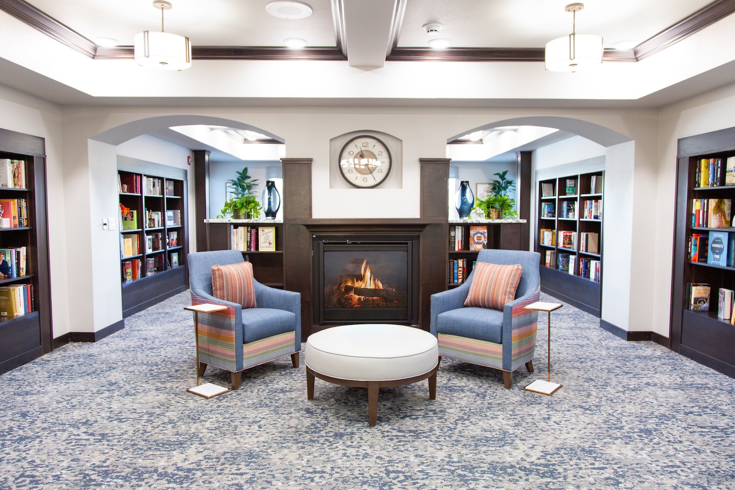 A cozy library room featuring two blue armchairs with striped cushions, a round white table between them, and a fireplace. Bookshelves filled with books line the walls, and a clock hangs above the fireplace. Warm lighting adds to the ambiance.