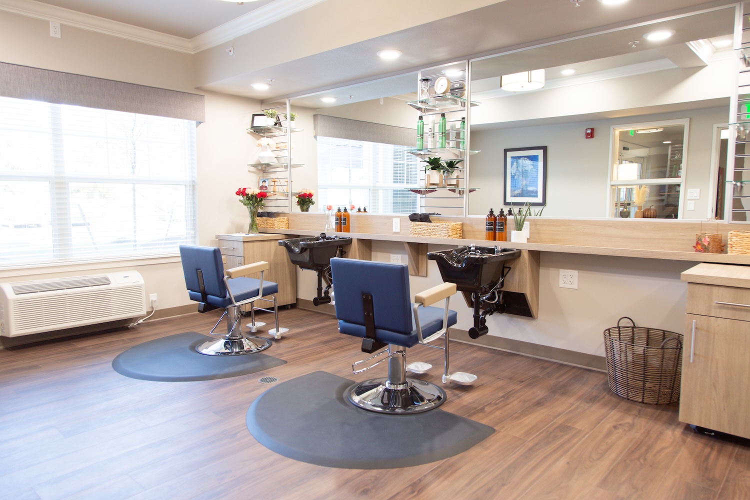 Bright, modern hair salon interior with two blue salon chairs facing large mirrors. Shelves hold hair products, and large windows let in natural light. Brown wood flooring and neutral walls create a welcoming atmosphere.