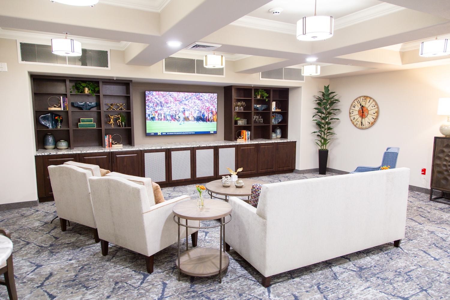 A modern living room with two white sofas facing a wall-mounted TV displaying a sports game. Dark wooden shelves with decor flank the TV. A large clock decorates the wall, and several hanging lights illuminate the space.