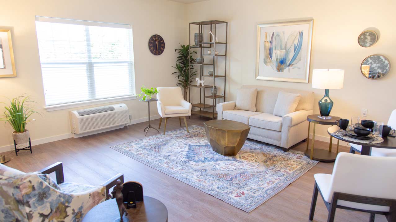 A cozy living room with a large window, light-colored walls, and a patterned rug. It features a beige sofa, white armchairs, a unique brass coffee table, a stylish bookshelf, and framed artwork. A plant and various decor items complete the space.