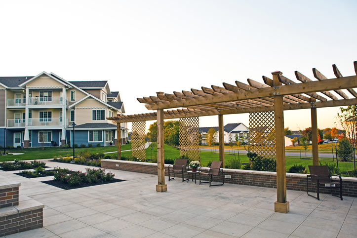 An outdoor patio area with a wooden trellis and lattice structure designed for senior living. The space includes several chairs and tables. In the background, there's a multi-story residential building with balconies and a green lawn, all bathed in the warm light of the setting sun.
