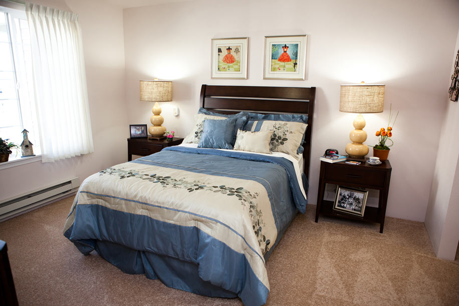 A cozy bedroom in a senior living residence features a neatly made bed with blue and white bedding, flanked by two dark wood nightstands with matching lamps. Above the bed hang two framed pieces of artwork. A window with vertical blinds allows light to brighten the room.