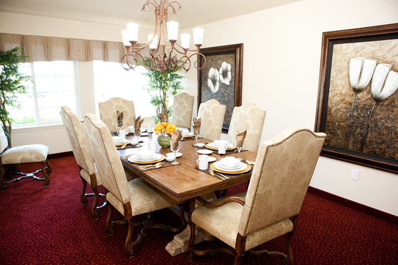 A dining room in a senior living community with a large wooden table set for eight, featuring white upholstered chairs. The table is adorned with plates, glasses, and a centerpiece of flowers and fruit. The room has a red carpet and framed floral artwork on the walls. A chandelier hangs above.