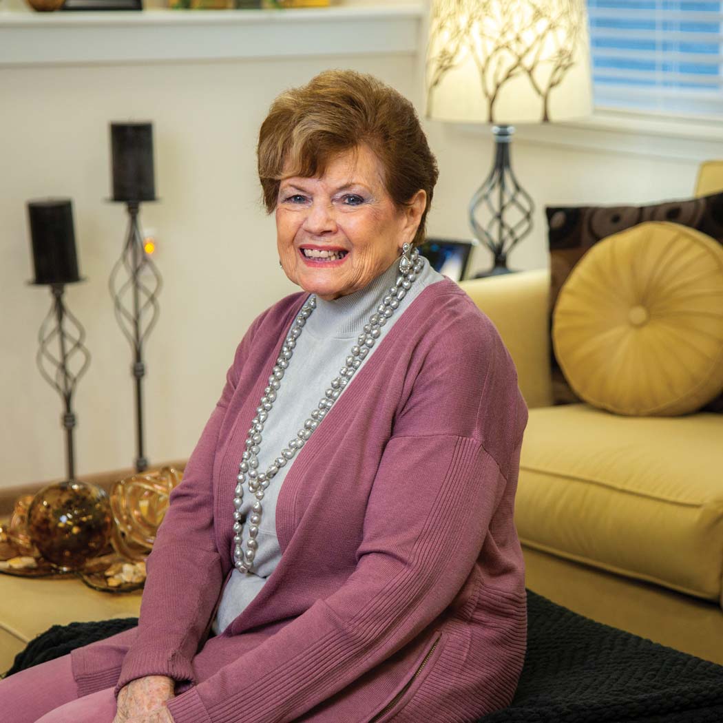 An elderly woman with short brown hair and a warm smile is sitting on a couch in her cozy Independent Living apartment. She is wearing a mauve cardigan, a gray turtleneck, and a long pearl necklace. Behind her are decorative lamps and candles on a white shelf.