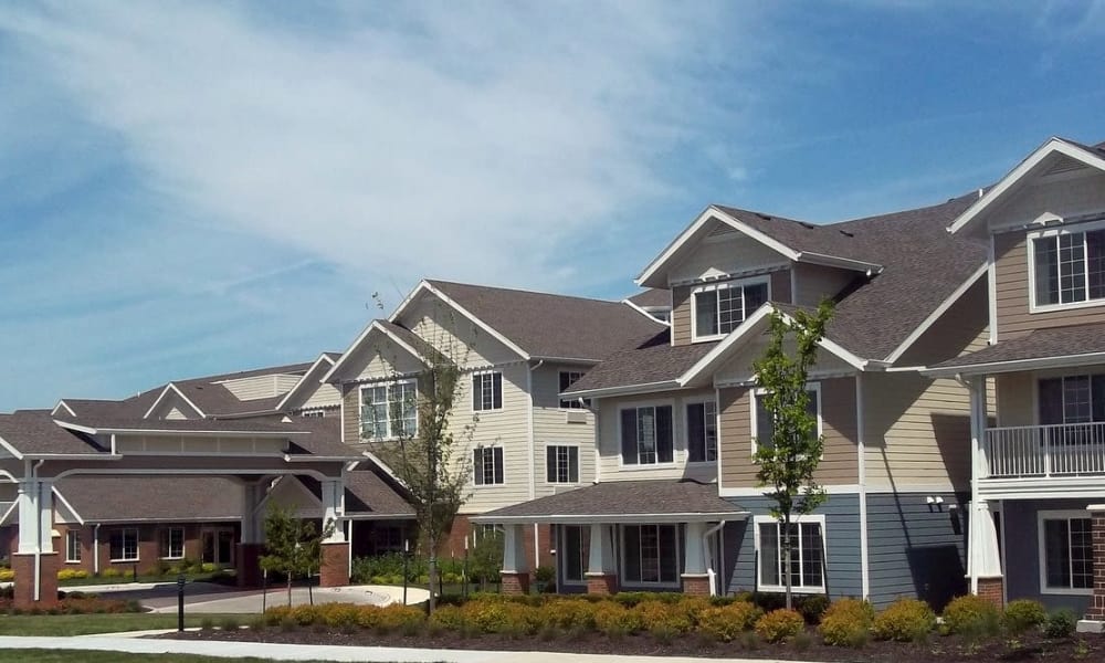 A modern senior living retirement community complex featuring multiple two-story buildings with beige and brown siding. The buildings are surrounded by well-kept landscaping with green vegetation and a clear blue sky overhead.