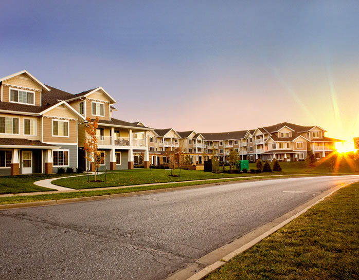 A peaceful suburban street at sunset features spacious, multi-story residential buildings with balconies. The warm light of the setting sun casts a golden glow over the well-maintained lawns and clear, calm road.