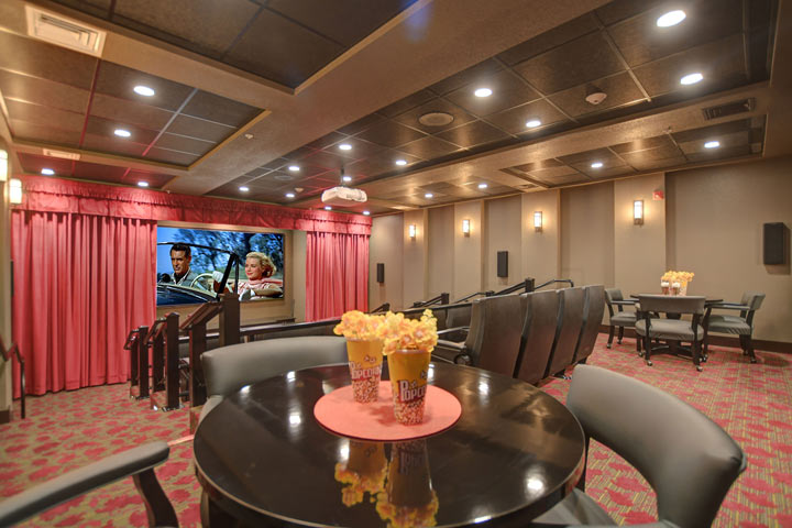 A private home theater with red carpeting and walls features a large screen displaying a movie scene with a man and woman in a car. Several tiered rows of leather reclining seats and small tables with popcorn create a cozy and cinematic atmosphere.