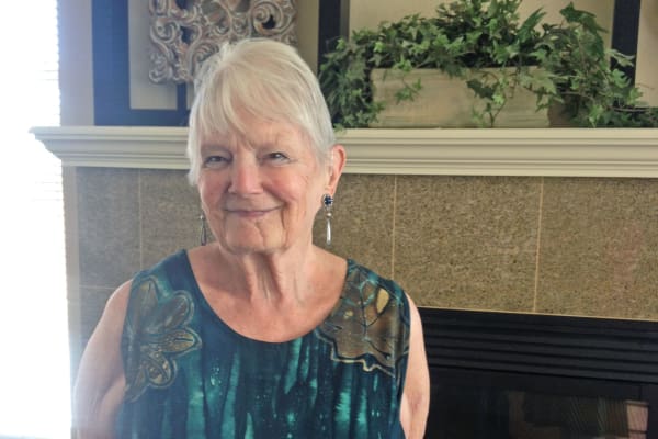 An elderly woman with short white hair stands in front of a fireplace, smiling. She is wearing a sleeveless, patterned dress and dangling earrings. The mantle above the fireplace is decorated with a potted plant and other ornamental items.