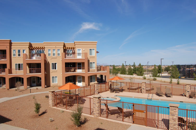 A modern apartment complex with beige and orange tones, featuring multiple balconies. In the foreground, there is a fenced outdoor pool area with lounge chairs and orange umbrellas. The surrounding area is landscaped with rocks and small plants under a clear blue sky.