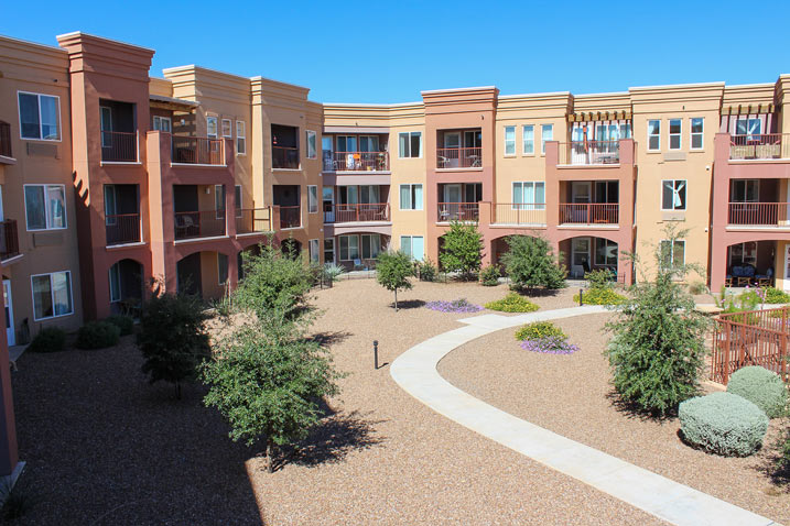 A courtyard view of a modern, three-story apartment complex with terra-cotta-colored walls and numerous balconies. The landscaped courtyard features gravel paths, small trees, and shrubbery against a clear blue sky.