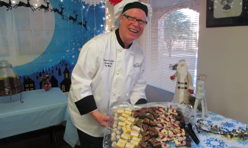 A smiling chef wearing a white coat and black hat holds a large platter of assorted desserts, including brownies and lemon squares. The background features festive holiday decorations, such as a Santa Claus figure and a winter-themed backdrop.