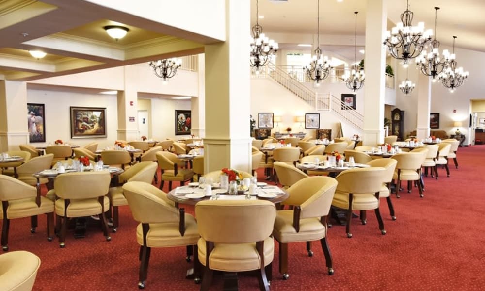 A spacious, well-lit dining area with elegant chandeliers, red carpet flooring, and numerous round tables surrounded by beige upholstered chairs. The tables are set with white tablecloths, silverware, and small floral centerpieces. Large windows and artwork adorn the walls.