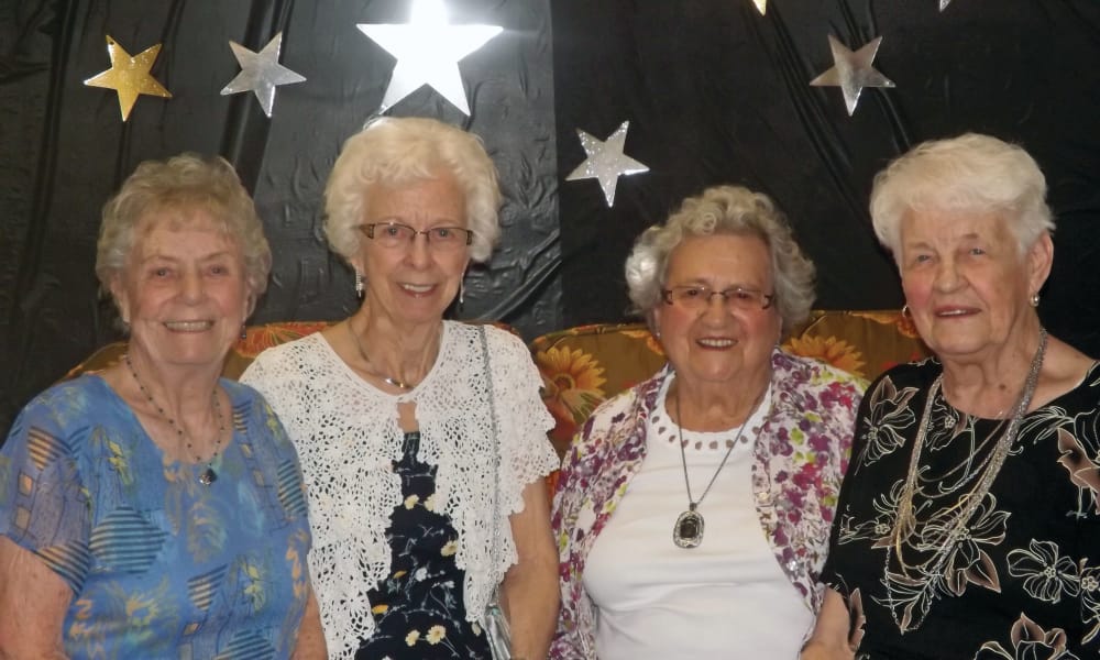 Four elderly women are standing together, smiling at the camera against a backdrop decorated with stars. They are dressed in various patterned tops and jewelry, creating a festive and joyful atmosphere.
