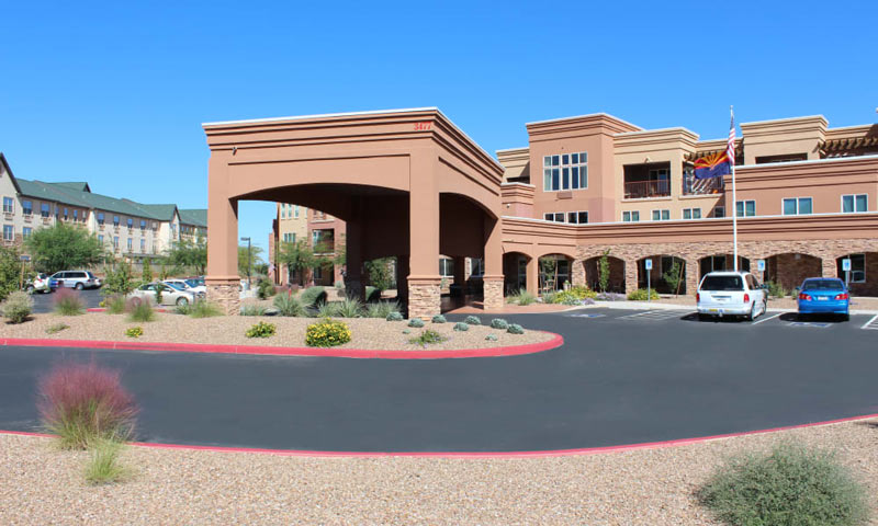 A modern brick building with a covered entrance, surrounded by a landscaped area with gravel and desert plants. Cars are parked near the entrance, and flags are visible near the building. The sky is clear and blue.