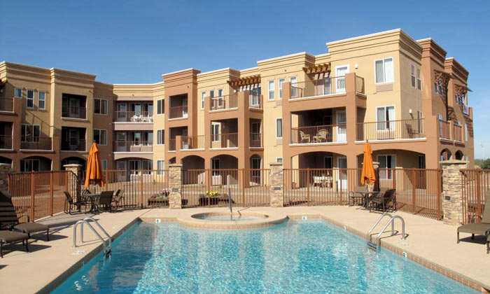 A modern apartment complex with three floors, featuring a central swimming pool surrounded by lounge chairs and umbrellas. The buildings have balconies and a beige and brown color scheme. The sky is clear and blue.